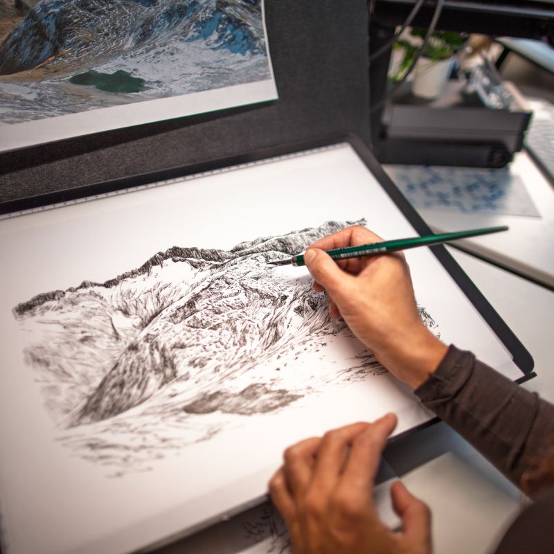 Artist working on an intricate Alpine mountain illustration at a drawing desk
